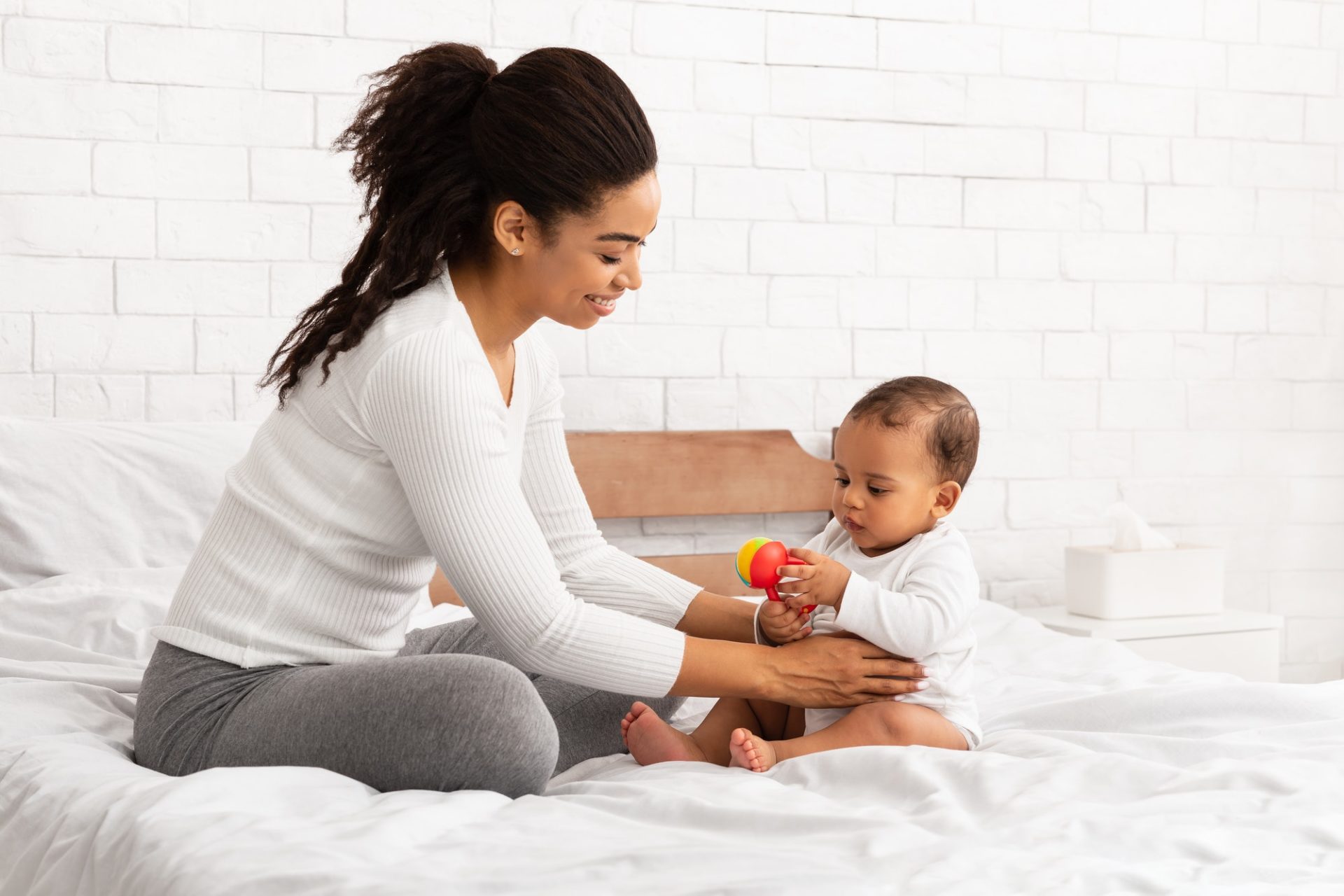 black-mother-playing-with-baby-giving-toy-sitting-in-bedroom-e1634190416345.jpg